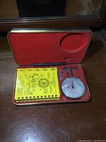 Diamond gauge kit in open metal case showing dial gauge, metal gauge, and yellow instruction booklet.