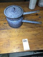 Old enamel ware double boiler pot with speckled grey and white finish on wooden table, showing side view of lid and handles.