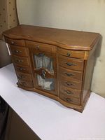 Front view of wooden musical jewelry box showing multiple drawers and a central glass display cabinet with decorative metal handles.