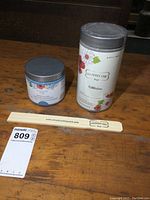 Two jars of unopened Country Chic paint and a branded wooden stir stick on a wooden surface. The large jar is white packaging with floral design, the small jar is blue labeled Tide Pool.