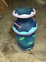 Front angle showing the blue metal milk can with green leaf designs and purple handles, with scalloped lid on top.