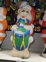 Front and side views of a vintage plastic teddy bear blow mold figure playing a drum, showing detailed costume and drum features with attached power cord.