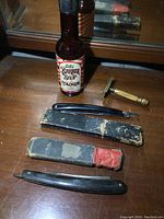 Full shot showing two vintage straight razors with black handles, two worn cases and a brown glass bottle of Fuller Barber Shop Cologne on a wooden surface.