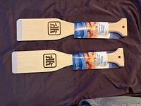 Two identical wooden BBQ scrapers with flat scraping edges and ergonomic handles laid out on a brown surface, showing front side with branding and label tags.