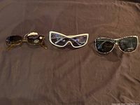 Three pairs of sunglasses laid flat on a brown surface showing their front view: tortoiseshell oval frame, white wraparound frame, and black cat-eye frame.