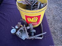 Yellow bucket filled with a miscellaneous assortment of metal rollers and brackets on a table outdoors, showing rust and wear.