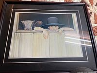 Framed print showing a boy in a black hat and a cow looking over a pale wooden fence, photo with reflection from lighting visible on glass.