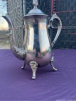Front side view showing the full coffee pot standing on four decorative feet, with ornate handle and spout details, and closed lid.