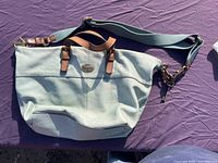 Light blue leather purse with tan handles and adjustable shoulder strap laid flat on purple cloth