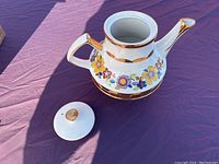Teapot with removed lid showing flower pattern and gold accents on handle and spout, sitting on purple cloth.