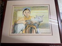 Framed print showing girl holding a white lamb, entire artwork and frame visible, lighting highlights colors and texture of the print.