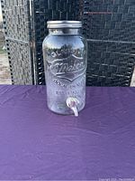 Clear large glass mason jar beverage dispenser with metal lid and spigot on purple surface in front of wicker background