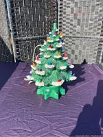 Photo showing vintage ceramic Christmas tree with multicoloured bulbs, green with white snow accents, placed on purple cloth background.