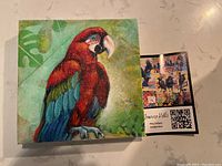 Front view of the square 8x8 parrot painting on canvas with vibrant colours and background leaf designs, accompanied by a business card showing the artist's details.