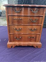 Front view of a compact wooden jewelry box with four drawers, brass-tone handles, scalloped base, placed on a purple cloth.