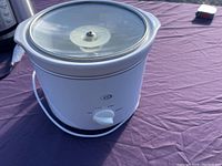 White small slow cooker with a round glass lid on a purple surface, showing front dial control with Off, Low, and High settings and attached power cord.