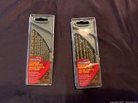 Photo of two packs of Benchmark brand titanium drill bit sets, each containing 7 pieces in clear plastic packaging with red and yellow labels.