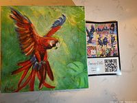 Square 8x8 canvas painting depicting a red and blue parrot in flight against a green background, with an artist's promotional card next to it.
