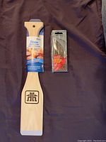 Wood BBQ scraper with label attached and titanium drill bit set in original transparent packaging displayed on fabric background.