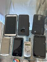 Image showing six cellphones/tablets including white Samsung phone, black HTC phone, Apple iPhone, flip phone, and cases on glass table