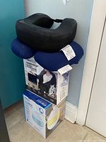 Four items stacked: two neck pillows (black and blue) on top, boxed Homedic heated wrap below, and boxed humidifier at bottom.
