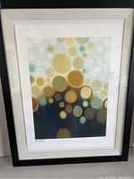 Full view of the framed abstract art print featuring gold, yellow, and teal circular shapes with white matting and black frame.