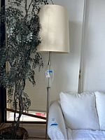 Tall floor lamp standing next to a window and couch, showing full height and fabric shade.