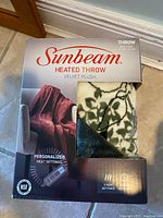 Photo of Sunbeam heated throw packaging showing velvet plush throw with green leaf design and features like 3 heat settings and personalized controls.