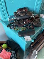 Pile of multiple sunglasses stacked on a teal cabinet with several hard cases visible. Various frame styles include tortoiseshell and black plastic. Photos focus on the condition and variety of sunglasses in the lot.