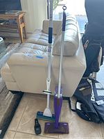 Photo showing two Swiffer sweepers with handles standing upright against a white couch, one in purple and one in white and green, and a black item on the floor nearby.