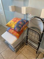 Photo of four colorful decorative pillows stacked on two light-colored elevated back pillows, resting on a grey folding chair next to a silver metal towel rack