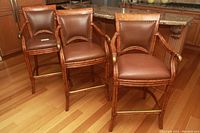 Front view of three identical barstools placed at kitchen counter showing leather seats with nailhead trim, wooden frames, armrests, and metal footrests.