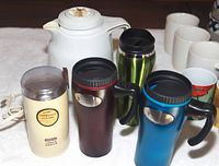Photo of a coffee collection with a white Thermos coffee pot, three metallic insulated mugs in red, blue, and green, each with lids and handles, and a Waring coffee grinder in off-white.