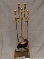 Photo of five-piece brass fireplace tool set on stand, showing broom, shovel, clamp, pike and mesh base stand