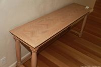 Long rectangular side table with a chevron wood pattern on the tabletop and turned legs, placed on a wooden floor against a wall.