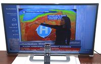 Front view of the Vizio 32 inch TV powered on, displaying weather graphics with remote control standing in front of the TV.