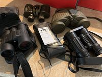 Overview of five binoculars arranged on surface including two olive green pairs, two black models, Minox in case