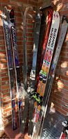 Photo showing seven pairs of downhill skis of various colors leaning against a brick wall, some with visible bindings.
