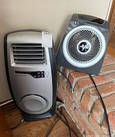 Vornado fan and Master Heat Plus heater positioned next to each other on wooden floor near brick fireplace.