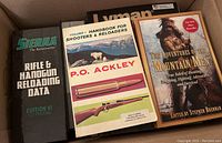 Box with multiple hardcover books about firearms, hunting, and reloading including Sierra Rifle & Handgun Reloading Data, Handbook for Shooters & Reloaders, and The Adventures of the Mountain Men.