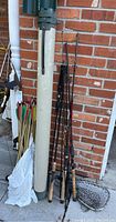 Full view showing cylindrical rod case along with grouped fishing rods leaning against brick wall and fishing net on pavement