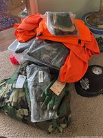 Pile of new military field jackets in camouflage and bright orange, new green wool mitts with leather fingertips, and wool blankets.