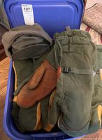 Blue container filled with winter and hunting gloves, mitts, and a hat. Gloves have olive green and brown leather sections with adjustable wrist straps. Gray and olive green hat is visible.