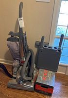 Full view showing Kirby Sentria upright vacuum with power cord, hose, dust bag and upright handle. Beside the vacuum is a grey plastic accessory holder standing on top of a Kirby shampoo system box.