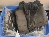 Six men's outerwear jackets in plastic bin with tags visible, olive and brown colors, folded inside bin.