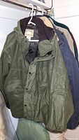 Photo showing green outdoor coat with hood, button closures, and front pockets hanging next to other jackets