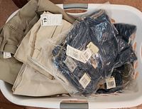 Laundry basket with folded tan and beige pants, some with tags, plastic sealed dark denim jeans from Columbia and Cole Haan