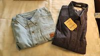 Two folded shirts side by side on beige surface, one light blue denim with L.L.Bean label visible, other dark heathered with Savane tag
