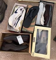 Five pairs of men's shoes and sandals including white Nike sneakers, black Teva sandals, brown Skechers loafers, placed in boxes on floor.