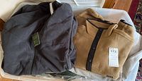 View of folded dark-colored zip-up jacket with tag and tan pullover fleece with black snaps along with other items in plastic bin.
